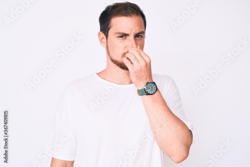 Young handsome man wearing casual white tshirt smelling something stinky and disgusting, intolerable smell, holding breath with fingers on nose. bad smell