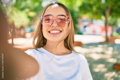 Young beautiful blonde caucasian woman smiling happy outdoors on a sunny day wearing heart shaped sunglasses taking a selfie picture