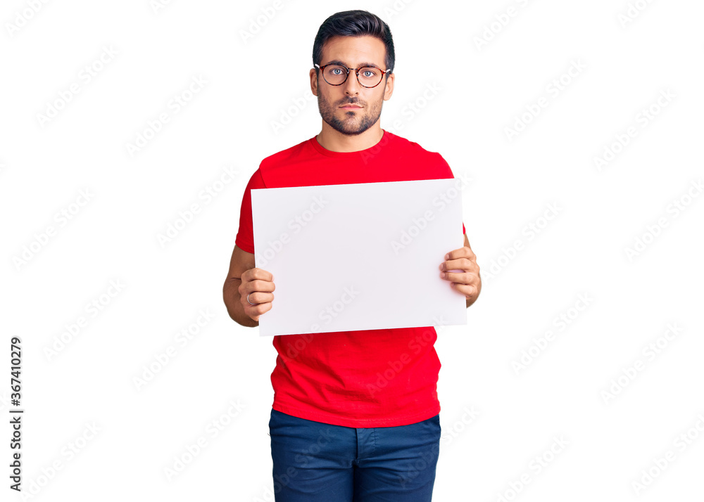 Young hispanic man holding blank empty banner thinking attitude and sober expression looking self confident Young hispanic man holding blank empty banner thinking attitude and sober expression looking self confident