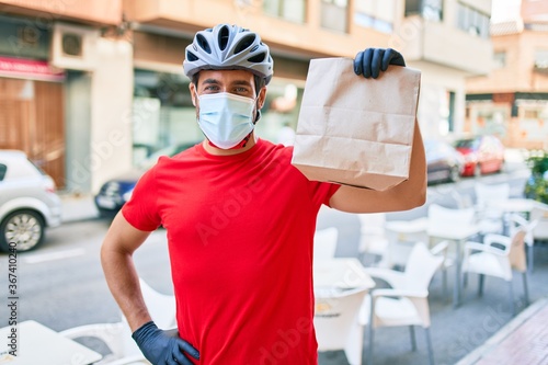 Young delivery man wearing bike helmet and coronavirus protection medical mask holding take away paper bag at town street.