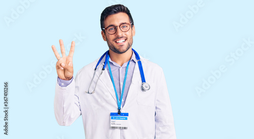 Young hispanic man wearing doctor uniform and stethoscope showing and pointing up with fingers number three while smiling confident and happy.