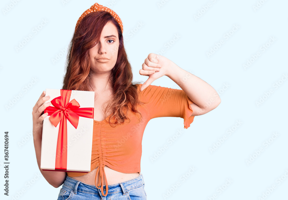 Young beautiful woman holding gift with angry face, negative sign showing dislike with thumbs down, rejection concept Young beautiful woman holding gift with angry face, negative sign showing dislike with thumbs down, rejection concept