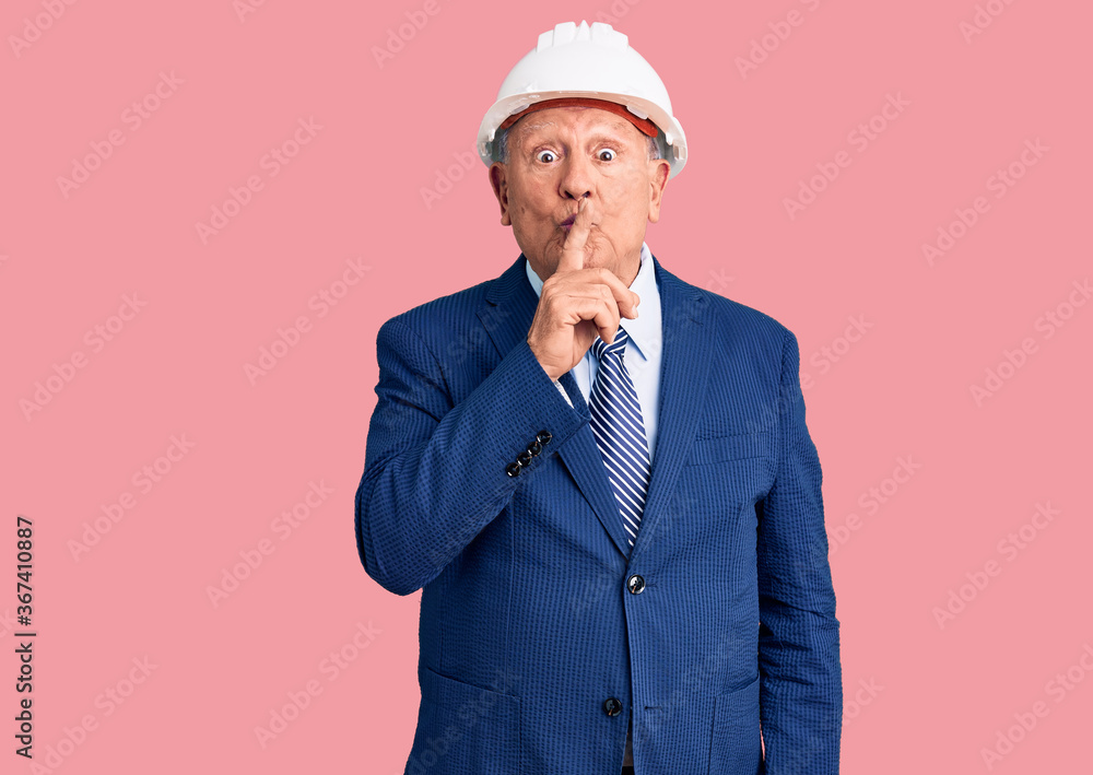 Senior handsome grey-haired man wearing suit and architect hardhat asking to be quiet with finger on lips. silence and secret concept.