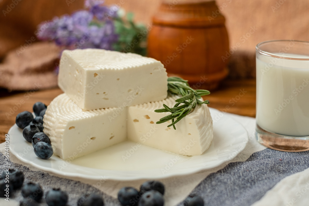 A head of cheese in a white plate lies on the table with blueberries and rosemary. Fresh goat cheese lies on a wooden table.