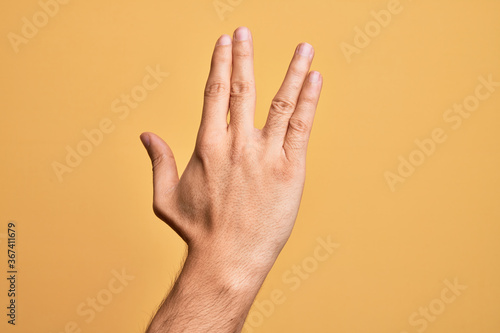 Hand of caucasian young man showing fingers over isolated yellow background greeting doing Vulcan salute, showing back of the hand and fingers, freak culture