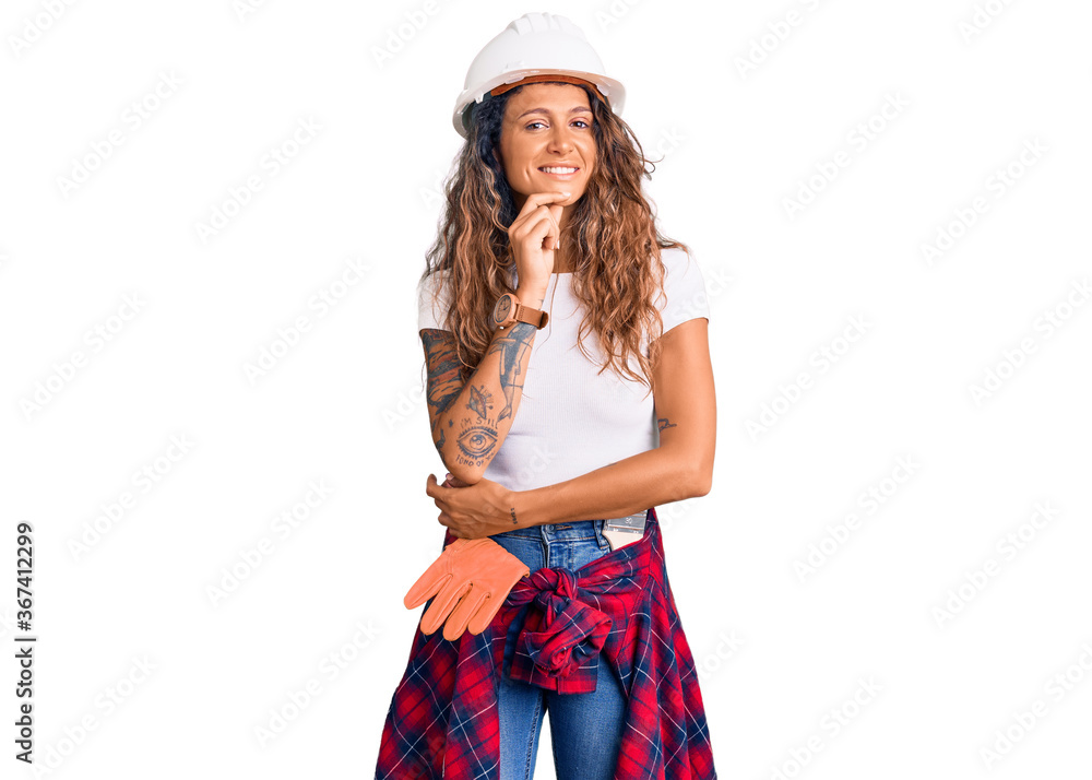 Young hispanic woman with tattoo wearing hardhat and builder clothes looking confident at the camera with smile with crossed arms and hand raised on chin. thinking positive. Young hispanic woman with tattoo wearing hardhat and builder clothes looking confident at the camera with smile with crossed arms and hand raised on chin. thinking positive.