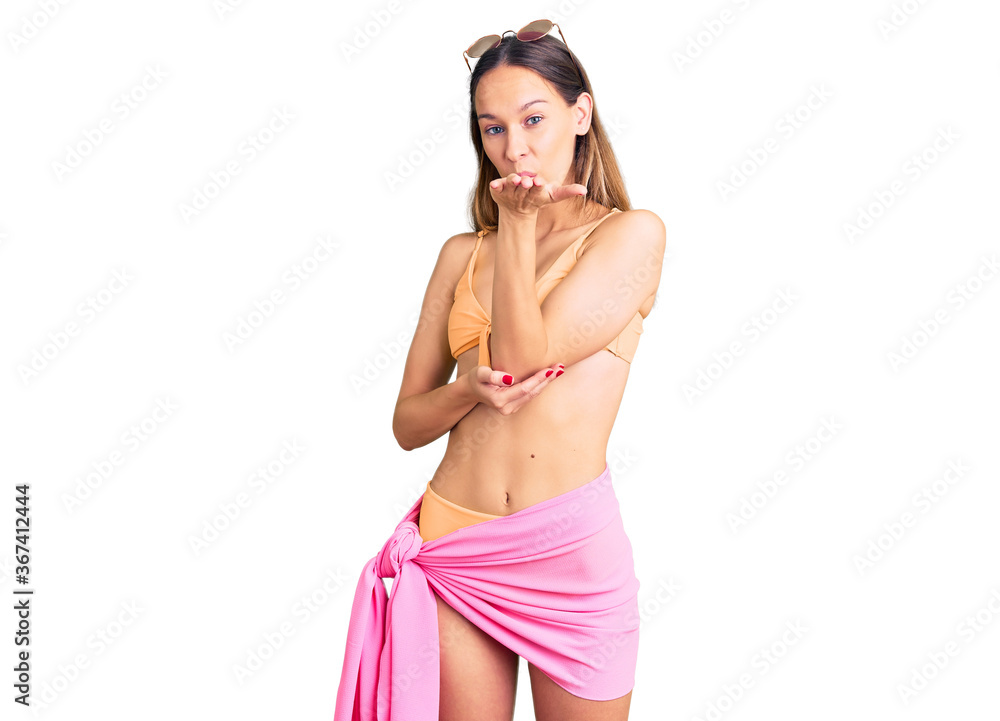 Beautiful brunette young woman wearing bikini and sunglasses looking at the camera blowing a kiss with hand on air being lovely and sexy. love expression. Beautiful brunette young woman wearing bikini and sunglasses looking at the camera blowing a kiss with hand on air being lovely and sexy. love expression.