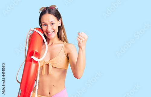 Beautiful brunette young woman wearing bikini and holding lifesaver float screaming proud, celebrating victory and success very excited with raised arms