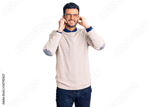 Young handsome hispanic man wearing elegant clothes and glasses covering ears with fingers with annoyed expression for the noise of loud music. deaf concept. © Krakenimages.com