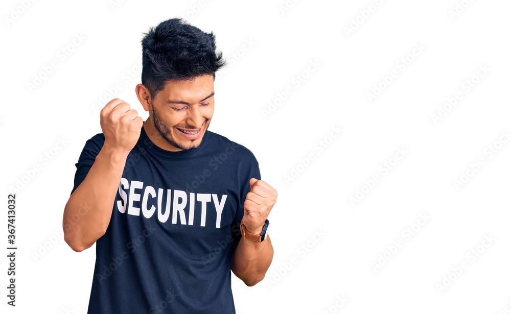 Handsome latin american young man wearing security t shirt very happy and excited doing winner gesture with arms raised, smiling and screaming for success. celebration concept. Handsome latin american young man wearing security t shirt very happy and excited doing winner gesture with arms raised, smiling and screaming for success. celebration concept.