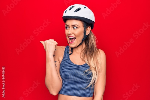 Young beautiful blonde cyclist woman wearing bike security helmet over red background pointing thumb up to the side smiling happy with open mouth