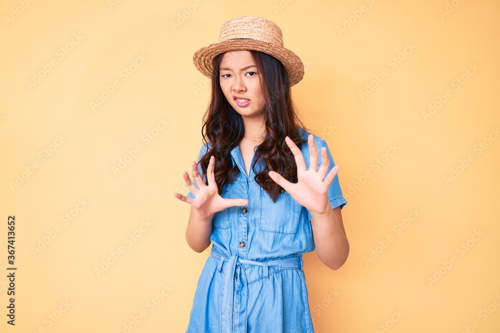 Young beautiful chinese girl wearing summer hat disgusted expression, displeased and fearful doing disgust face because aversion reaction. with hands raised Young beautiful chinese girl wearing summer hat disgusted expression, displeased and fearful doing disgust face because aversion reaction. with hands raised
