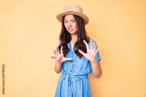 Young beautiful chinese girl wearing summer hat disgusted expression, displeased and fearful doing disgust face because aversion reaction. with hands raised