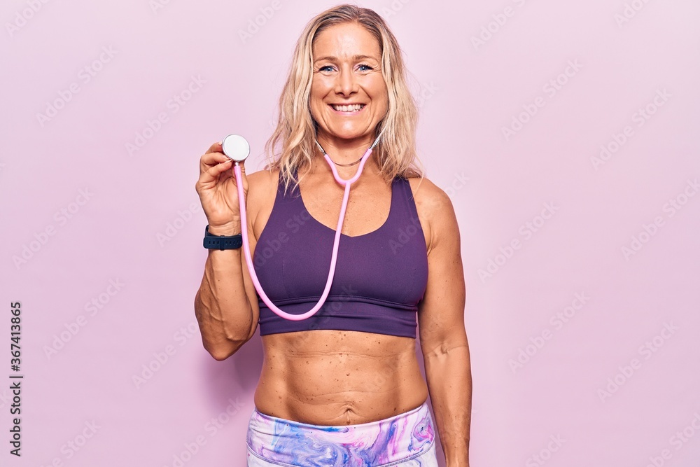 Middle age caucasian blonde woman wearing sport clothes holding stethoscope looking positive and happy standing and smiling with a confident smile showing teeth Middle age caucasian blonde woman wearing sport clothes holding stethoscope looking positive and happy standing and smiling with a confident smile showing teeth
