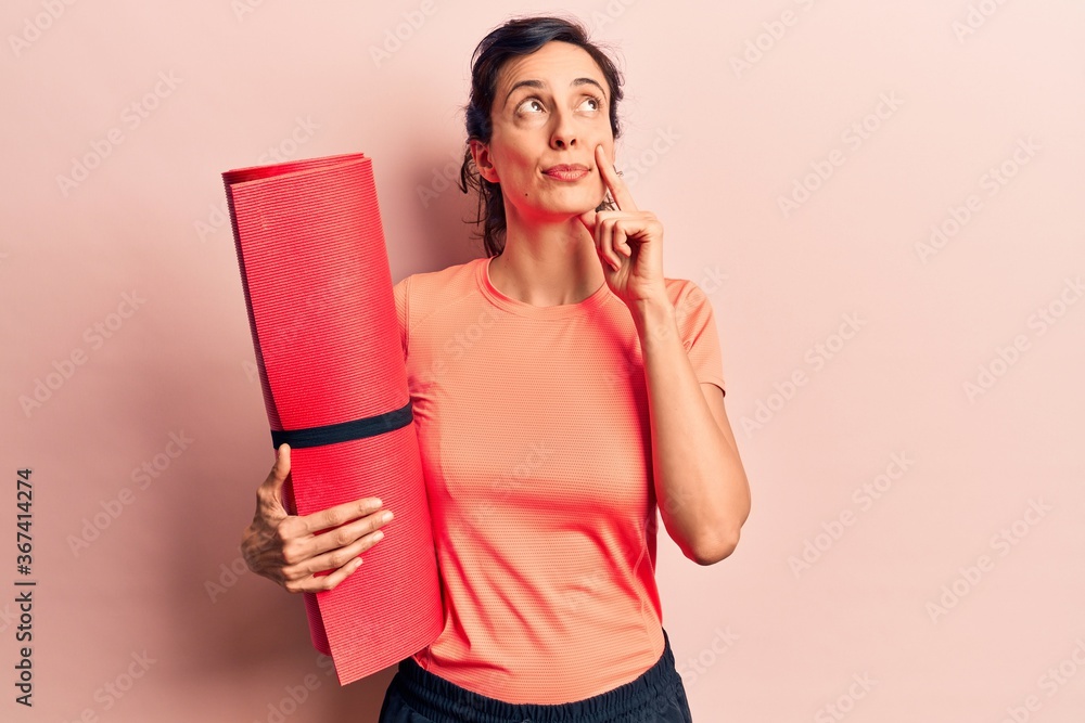 Young beautiful hispanic woman holding yoga mat serious face thinking about question with hand on chin, thoughtful about confusing idea Young beautiful hispanic woman holding yoga mat serious face thinking about question with hand on chin, thoughtful about confusing idea