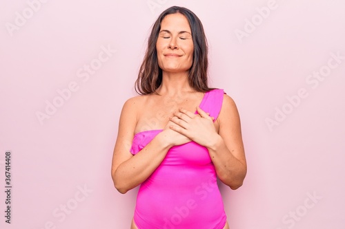Young beautiful brunette woman wearing pink swimwear standing over isolated background smiling with hands on chest  eyes closed with grateful gesture on face. Health concept.