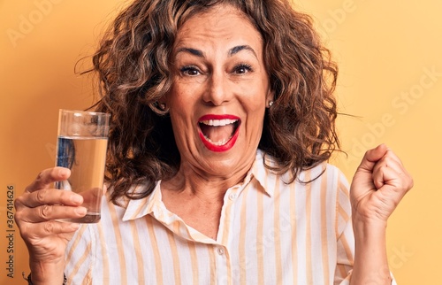 Middle age brunette woman drinking glass of water standing over isolated yellow background screaming proud, celebrating victory and success very excited with raised arm © Krakenimages.com