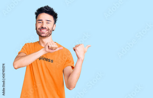 Young hispanic man wearing t shirt with happiness word message pointing to the back behind with hand and thumbs up, smiling confident