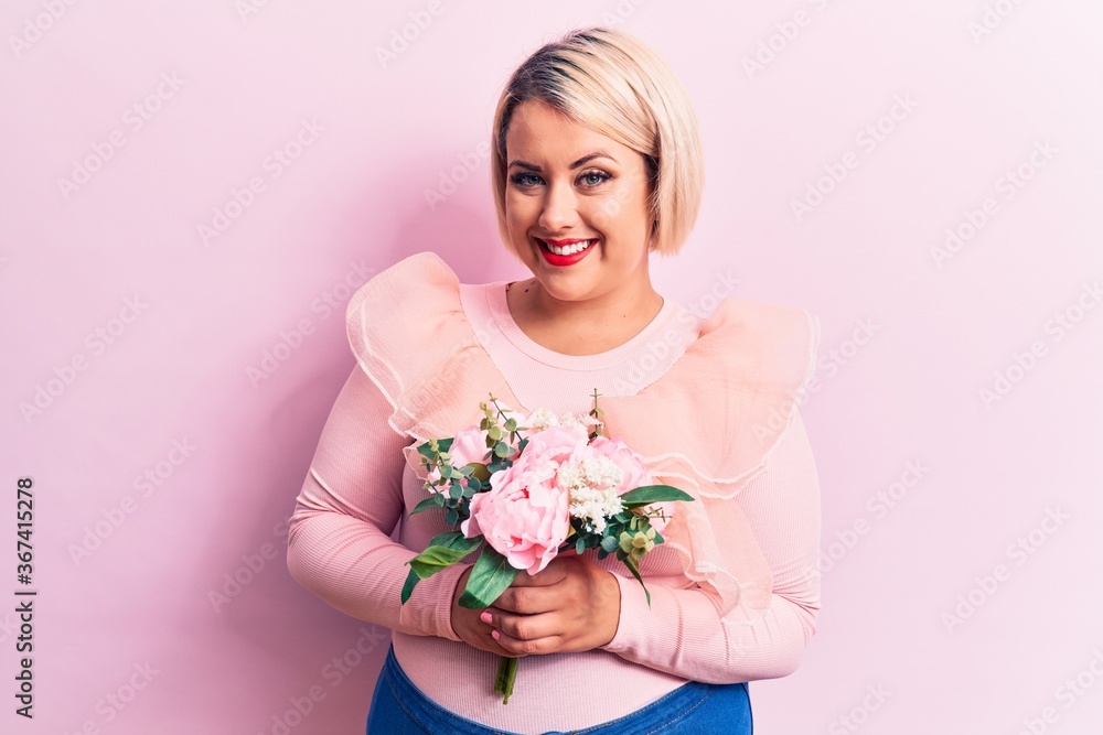 Beautiful blonde plus size woman holding bouquet of pink flowers over isolated background looking positive and happy standing and smiling with a confident smile showing teeth Beautiful blonde plus size woman holding bouquet of pink flowers over isolated background looking positive and happy standing and smiling with a confident smile showing teeth