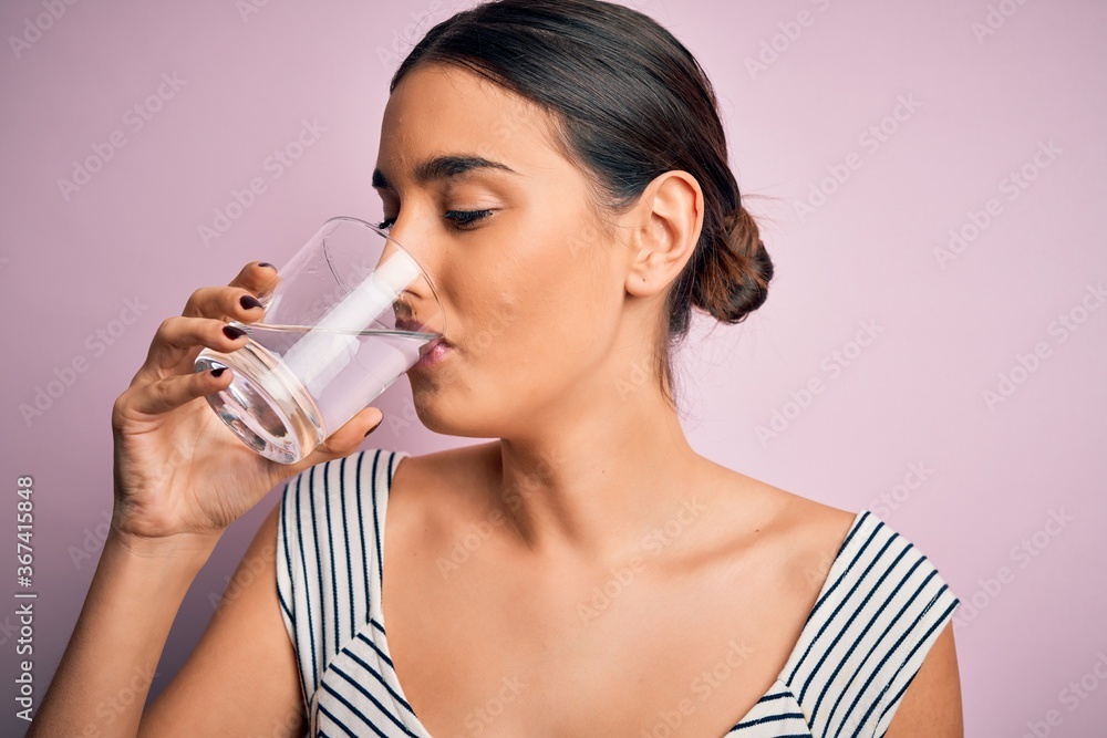 Young beautiful brunette woman drinking glass of healthy water to refreshment standing over isolated pink background Young beautiful brunette woman drinking glass of healthy water to refreshment standing over isolated pink background