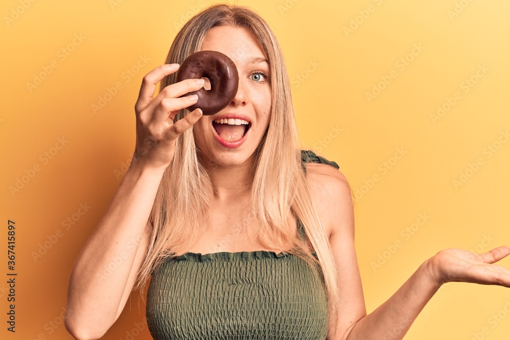 Fototapeta premium Young blonde woman holding donut celebrating achievement with happy smile and winner expression with raised hand