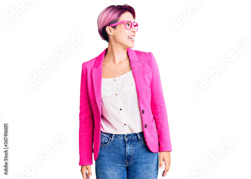 Young beautiful woman with pink hair wearing business jacket and glasses looking away to side with smile on face, natural expression. laughing confident. photo