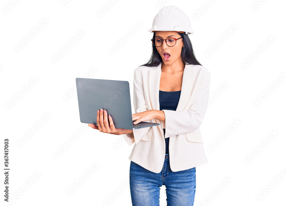 Young beautiful latin girl wearing architect hardhat holding laptop scared and amazed with open mouth for surprise, disbelief face Young beautiful latin girl wearing architect hardhat holding laptop scared and amazed with open mouth for surprise, disbelief face