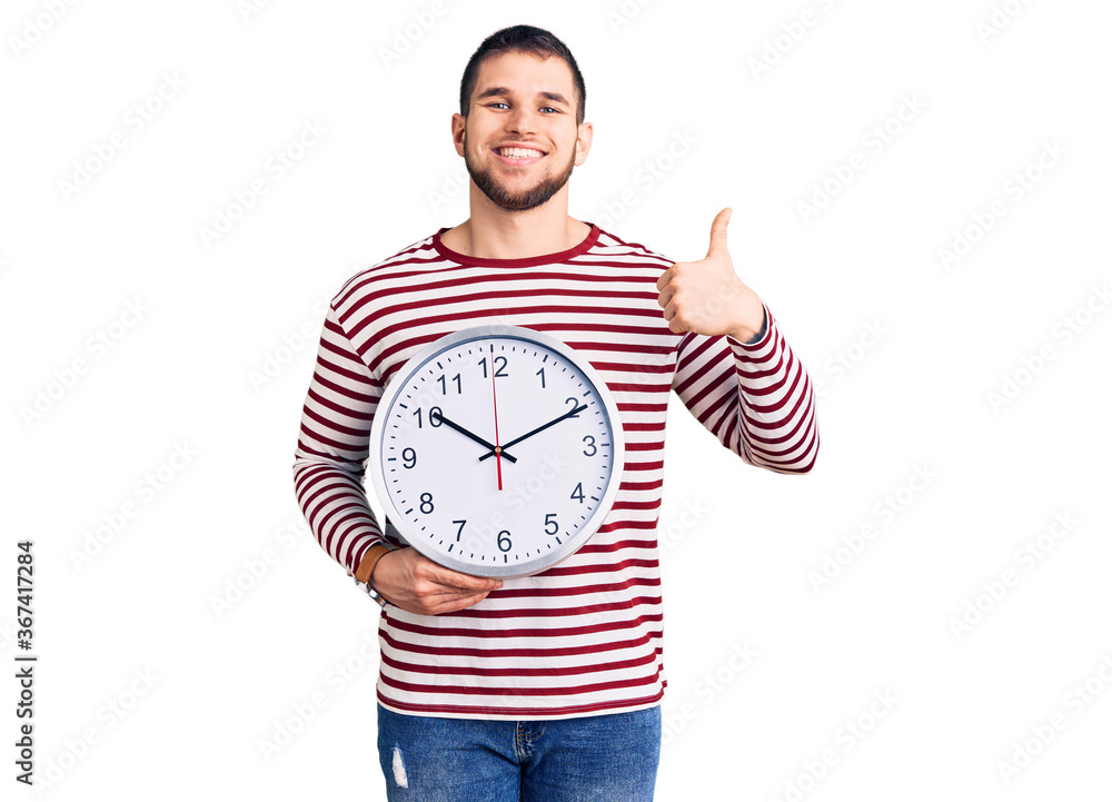 Young handsome man holding big clock smiling happy and positive, thumb up doing excellent and approval sign Young handsome man holding big clock smiling happy and positive, thumb up doing excellent and approval sign