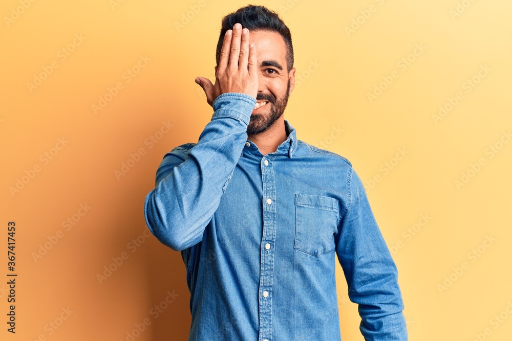 Fototapeta premium Young hispanic man wearing casual clothes covering one eye with hand, confident smile on face and surprise emotion.