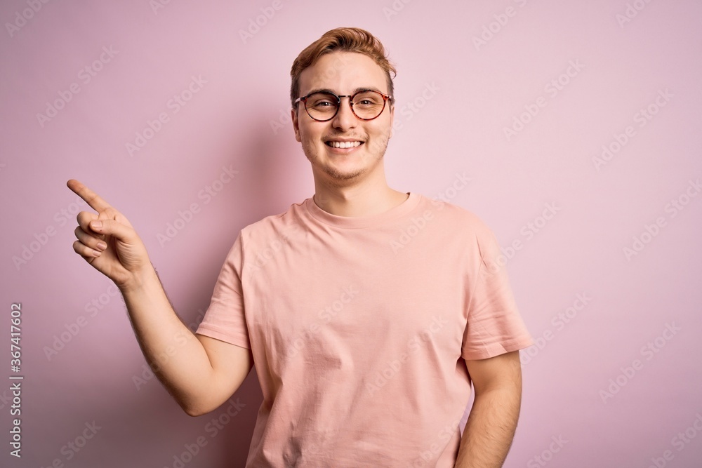 Young handsome redhead man wearing casual t-shirt standing over isolated pink background with a big smile on face, pointing with hand finger to the side looking at the camera.