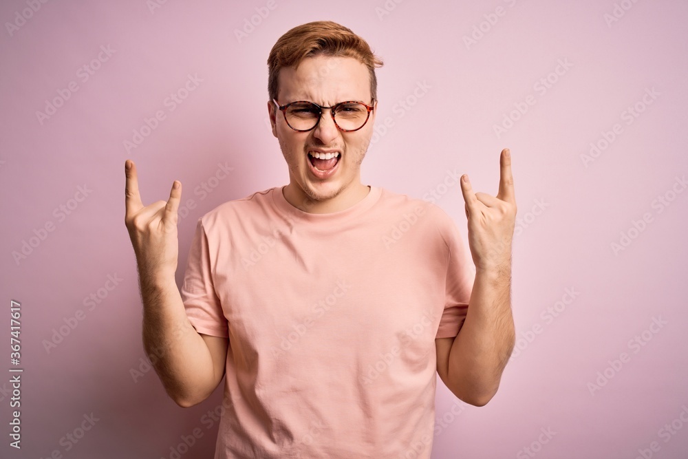 Young handsome redhead man wearing casual t-shirt standing over isolated pink background shouting with crazy expression doing rock symbol with hands up. Music star. Heavy music concept. Young handsome redhead man wearing casual t-shirt standing over isolated pink background shouting with crazy expression doing rock symbol with hands up. Music star. Heavy music concept.