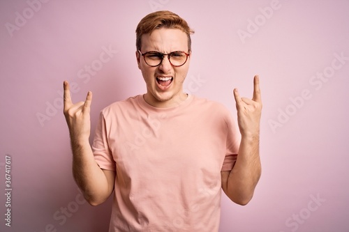 Young handsome redhead man wearing casual t-shirt standing over isolated pink background shouting with crazy expression doing rock symbol with hands up. Music star. Heavy music concept.