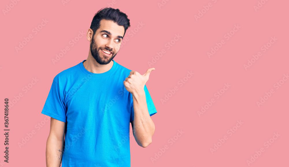 Young handsome man with beard wearing casual t-shirt smiling with happy face looking and pointing to the side with thumb up. Young handsome man with beard wearing casual t-shirt smiling with happy face looking and pointing to the side with thumb up.