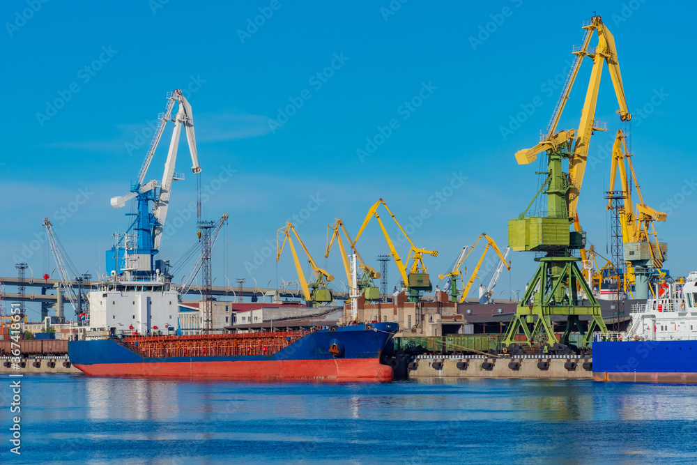 Seaport. Loading the ship. Red and blue ship at the dock in the port. Merchant fleet. The infrastructure of the commercial port. Port cranes against the blue sky.