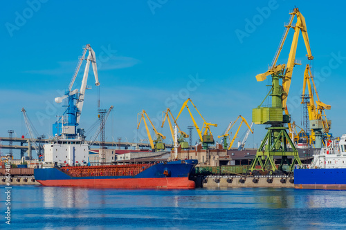 Seaport. Loading the ship. Red and blue ship at the dock in the port. Merchant fleet. The infrastructure of the commercial port. Port cranes against the blue sky.