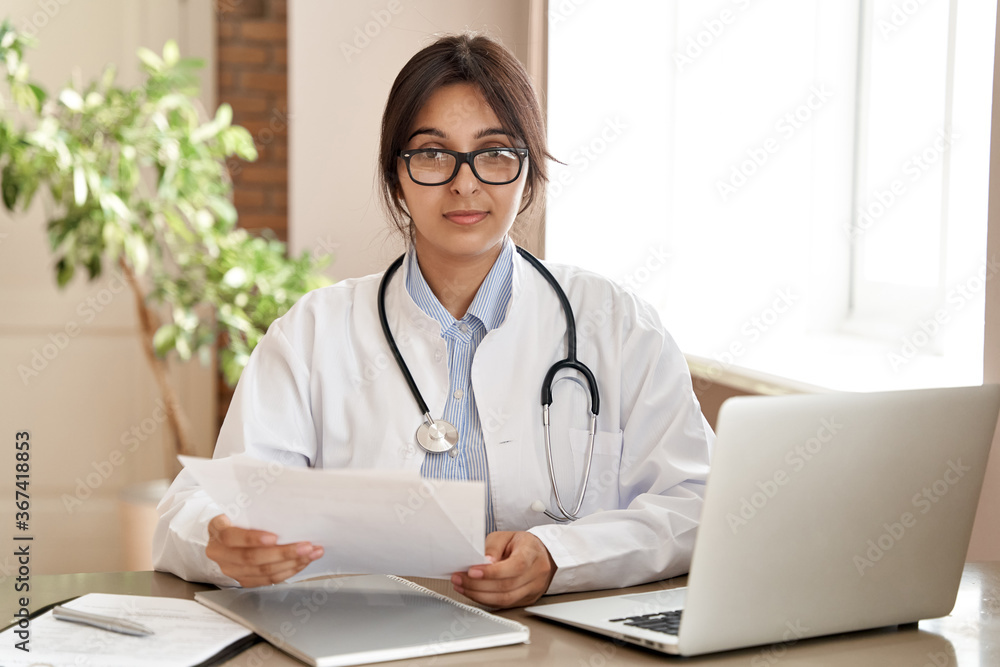 Confident indian female doctor wear white medical coat, glasses, stethoscope look at camera hold papers sit at workplace. Indian woman healthcare professional medic gp posing at work desk, portrait. Confident indian female doctor wear white medical coat, glasses, stethoscope look at camera hold papers sit at workplace. Indian woman healthcare professional medic gp posing at work desk, portrait.
