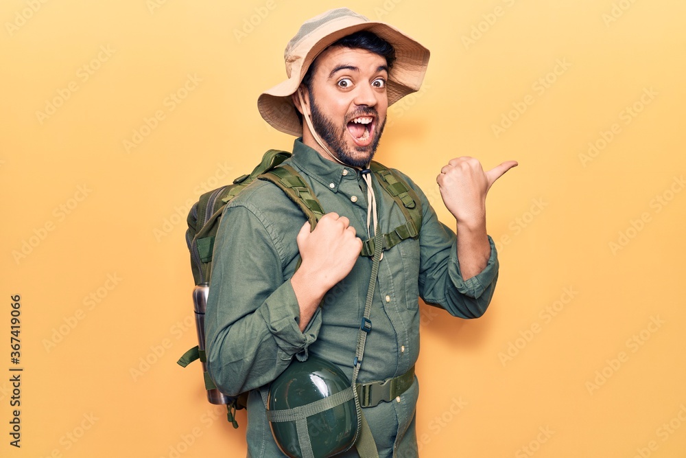 Young hispanic man wearing explorer hat and bakcpack pointing thumb up to the side smiling happy with open mouth Young hispanic man wearing explorer hat and bakcpack pointing thumb up to the side smiling happy with open mouth