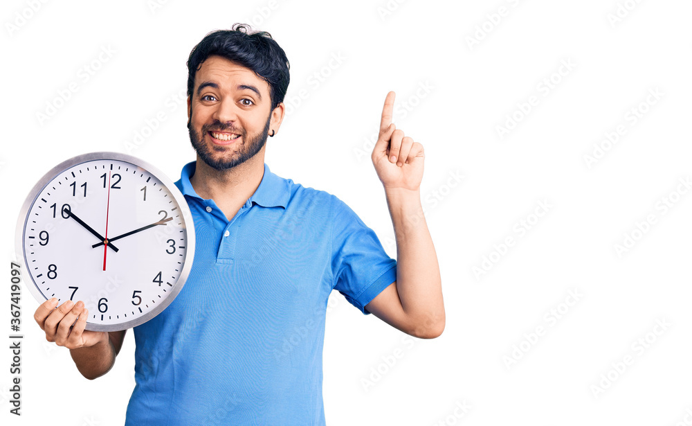 Young hispanic man holding big clock surprised with an idea or question pointing finger with happy face, number one Young hispanic man holding big clock surprised with an idea or question pointing finger with happy face, number one
