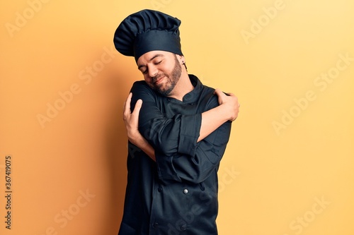 Young hispanic man wearing cooker uniform hugging oneself happy and positive, smiling confident. self love and self care