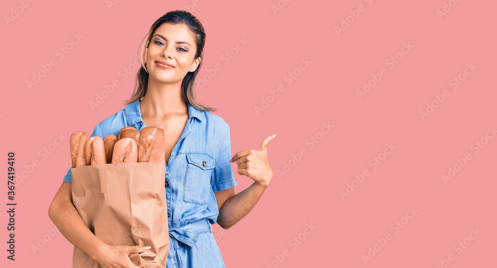 Young beautiful woman holding paper bag with bread pointing finger to one self smiling happy and proud Young beautiful woman holding paper bag with bread pointing finger to one self smiling happy and proud