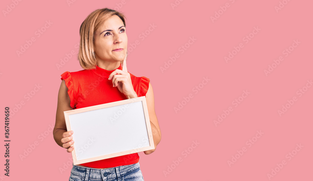 Young blonde woman holding empty white chalkboard serious face thinking about question with hand on chin, thoughtful about confusing idea Young blonde woman holding empty white chalkboard serious face thinking about question with hand on chin, thoughtful about confusing idea