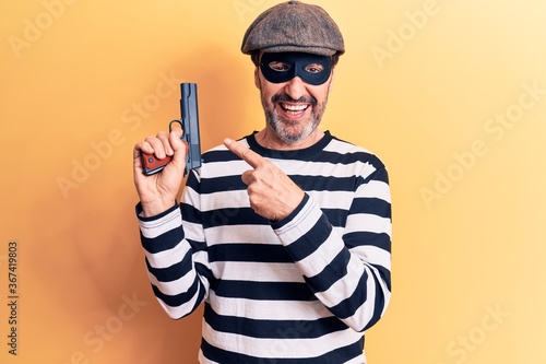 Middle age handsome burglar man wearing cap and mask holding gun over yellow background smiling happy pointing with hand and finger © Krakenimages.com
