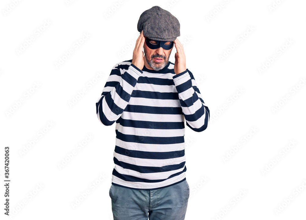 Middle age handsome man wearing burglar mask with hand on head for pain in head because stress. suffering migraine. Middle age handsome man wearing burglar mask with hand on head for pain in head because stress. suffering migraine.
