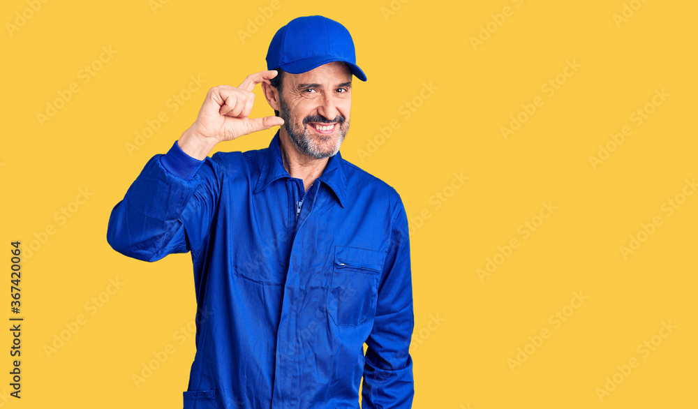 Middle age handsome man wearing mechanic uniform smiling and confident gesturing with hand doing small size sign with fingers looking and the camera. measure concept.