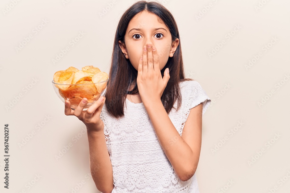 Beautiful child girl holding potato chip covering mouth with hand, shocked and afraid for mistake. surprised expression Beautiful child girl holding potato chip covering mouth with hand, shocked and afraid for mistake. surprised expression