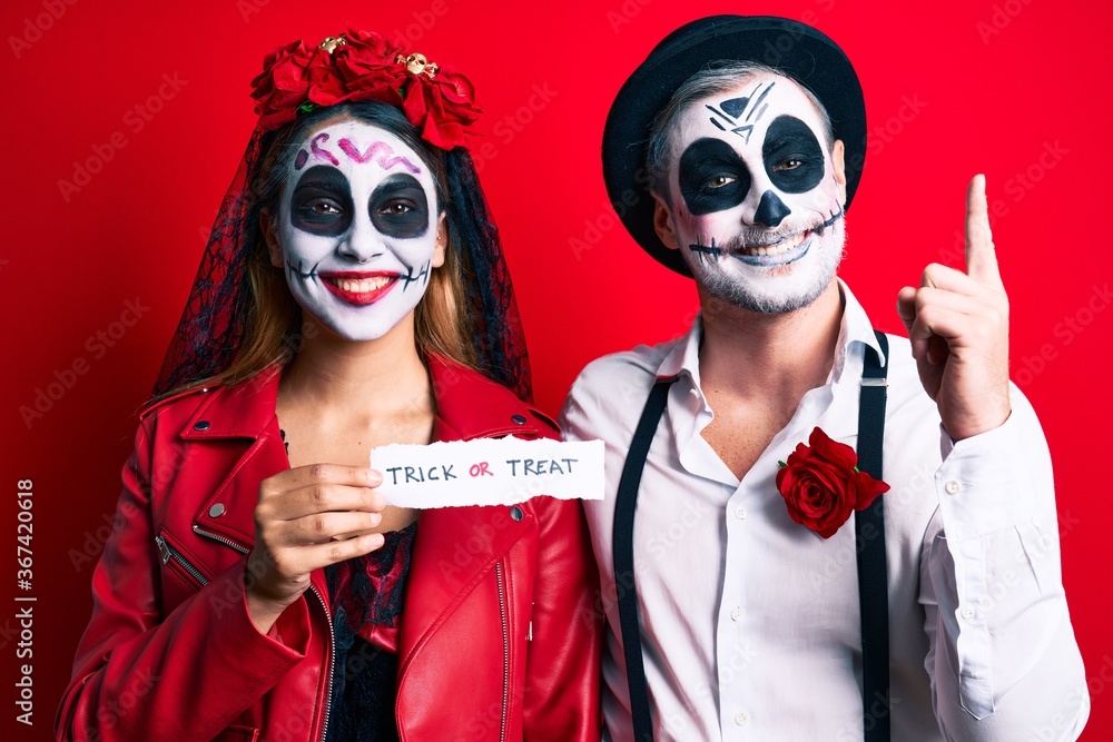 Couple wearing day of the dead costume holding trick or treat paper surprised with an idea or question pointing finger with happy face, number one Couple wearing day of the dead costume holding trick or treat paper surprised with an idea or question pointing finger with happy face, number one