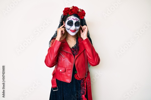 Woman wearing day of the dead costume over white covering ears with fingers with annoyed expression for the noise of loud music. deaf concept.