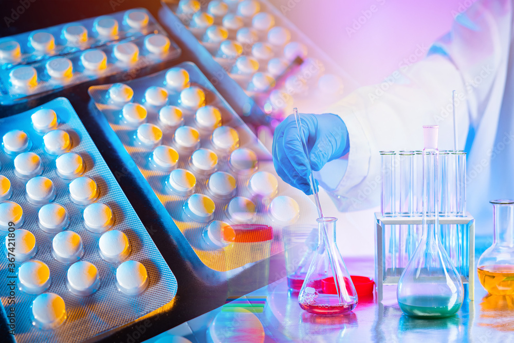 A table in a chemical laboratory and a chemist's hand on the background of blisters of tablets. Research in the field of medicine. Testing new drugs. Studying the properties of medicines.