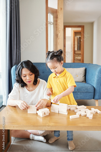 Young Asian woman and her little daughter playing with wooden bricks and making walls and towers
