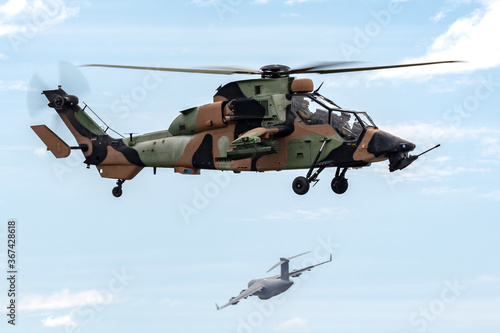 Armed reconnaissance helicopter hovering while a military cargo aircraft departs in the background.
